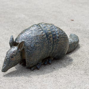 May include: A decorative armadillo figurine featuring a textured, segmented shell in shades of dark gray, blue, and brown. The sculpture displays a pointed snout, upright ears, and a long tail, capturing the animal in a low, resting posture.