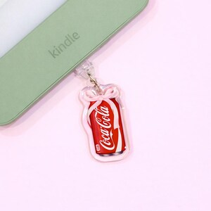 Puede incluir: Un llavero de lata de Coca-Cola roja y blanca con un diseño de lazo transparente. El llavero está sujeto a un dispositivo Kindle verde. El fondo es de color rosa claro.