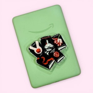 May include: A light green Amazon Kindle case with a stack of book-shaped pop sockets. The pop sockets feature book covers from the Twilight series, including "Twilight," "Eclipse," "Breaking Dawn," "Midnight Sun," and "New Moon."