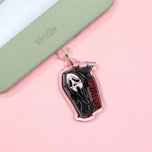Könnte beinhalten: Ein klarer Acryl-Anhänger mit einem schwarzen Sarg mit der Ghostface-Figur aus der Scream-Filmreihe. Der Anhänger hat eine Fledermaus oben und das Wort "GHOSTFACE" in Rot. Der Anhänger ist an einem Kindle befestigt.