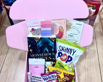 May include: A pink gift box filled with snacks, beauty products, and a book titled "Monstrous Urges." The box includes Skinny Pop popcorn, Welch's fruit snacks, and a lip balm.