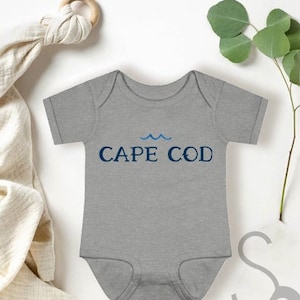 Puede incluir: Un body de bebé gris con mangas cortas y un gráfico de olas azul y blanco. El body tiene el texto "CAPE COD" impreso en la parte delantera.