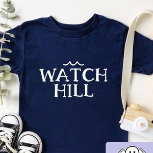 May include: A navy blue t-shirt with the text "WATCH HILL" in white lettering. The shirt is decorated with a wavy line above the text. The shirt is laid flat on a white surface with a pair of black and white sneakers and a wooden toy camera.