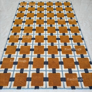 May include: A rectangular rug with a geometric pattern. The design features squares in shades of brown, navy blue, and light blue, arranged in a grid-like formation. The text "Kashi Rugs" is visible in several places on the rug.