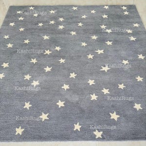 May include: A gray rug with a pattern of cream-colored stars scattered across its surface. The rug has the text "KashiRugs" printed on it in several places. The rug is a square shape.