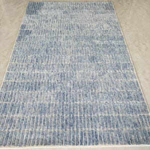 May include: A rectangular rug with a textured pattern in shades of blue and off-white. The design features vertical lines, creating a striped effect. The rug is placed on a light-coloured tile floor.