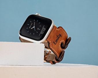 May include: A silver Apple Watch with a black screen displaying the time and date, paired with a brown leather band. The band is personalized with the name "Emily". The watch is displayed on a white surface against a blue background.