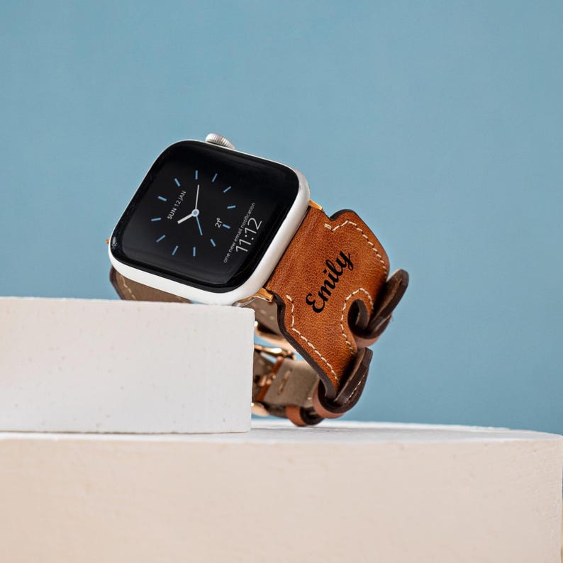 May include: A silver Apple Watch with a black screen displaying the time and date, paired with a brown leather band. The band is personalized with the name "Emily". The watch is displayed on a white surface against a blue background.