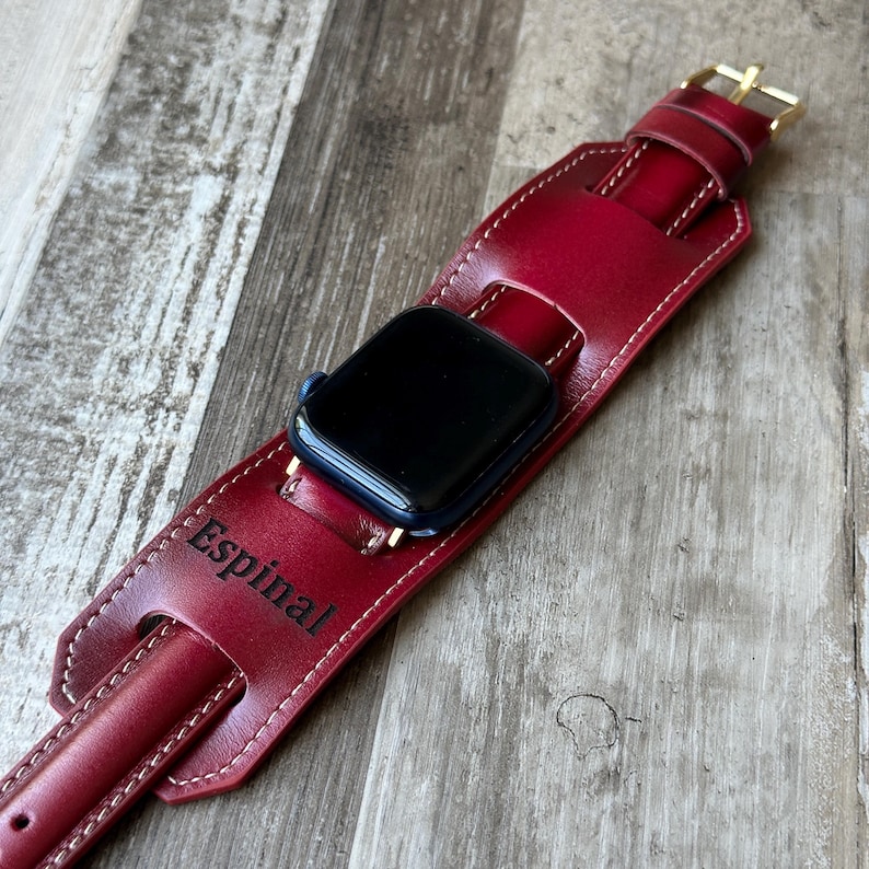 May include: A burgundy leather watch band with white stitching and a gold-colored buckle. The band holds a black smartwatch and has the name "Espinal" engraved on it. The watch band is on a wooden surface.