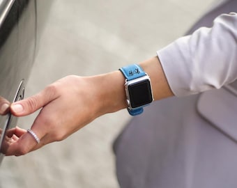 May include: A silver smartwatch with a light blue leather band. The band features a monogram in black. The watch is being worn on a wrist.