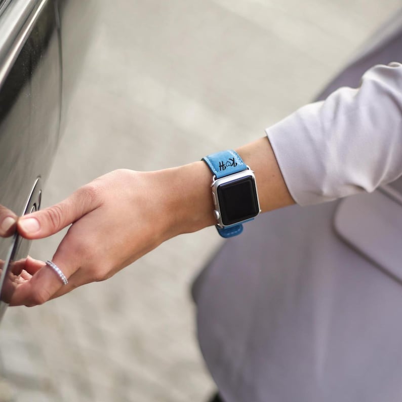 Peut inclure: Une montre connect&eacute;e argent&eacute;e avec un bracelet en cuir bleu clair. Le bracelet porte un monogramme noir. La montre est port&eacute;e au poignet.