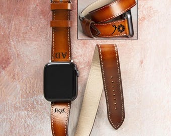 May include: Brown leather Apple Watch band with white stitching and a silver buckle. The band features personalized initials and a floral design. The watch face is black.