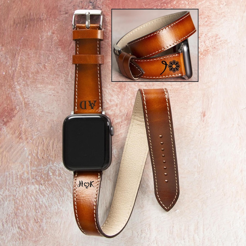 Puede incluir: Correa de reloj Apple de cuero marr&oacute;n con costuras blancas y hebilla plateada. La correa presenta iniciales personalizadas y un dise&ntilde;o floral. La esfera del reloj es negra.