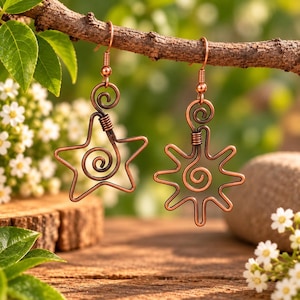 May include: A pair of copper-toned wire earrings hanging from a branch. One earring is shaped like a five-pointed star with a central spiral, and the other is shaped like a sun with multiple rays and a central spiral, both featuring hook wires.