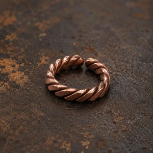 May include: A copper-colored ring featuring a twisted, rope-like design with a visible gap in the band. The metal surface has a textured, metallic finish with subtle variations in tone, creating a continuous spiral pattern around the circular band.