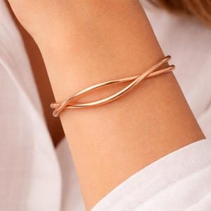 May include: A rose gold bracelet with a minimalist design. The bracelet features two curved, intersecting bands that create an open, elegant look. The jewellery is shown on a person's arm, against a white shirt.