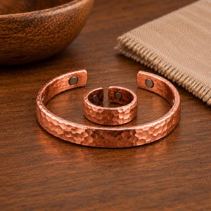 May include: A copper-coloured metal cuff bracelet and a matching open ring, both featuring a hammered texture on the exterior surface. Each piece includes small, circular dark-coloured magnets embedded on the interior ends of the metal bands.
