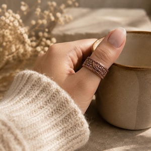 May include: A copper-colored ring with a floral and leaf pattern. The ring is worn on a thumb. The ring's design includes detailed floral and leaf patterns. The ring is a band style, and the metal has a warm, reddish-brown hue.
