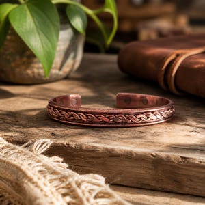 Op de afbeelding: Een koperkleurige manchetarmband met een gevlochten patroon. De open armband wordt gepresenteerd op een rustiek houten oppervlak. Het ingewikkelde ontwerp en de warme tinten van de armband creëren een vintage esthetiek. De armband is een sieraad.