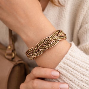 Puede incluir: Un brazalete trenzado en tonos cobre y latón. El brazalete presenta un diseño tejido con hebras alternas de cobre y latón. La joya se lleva en la muñeca, mostrando el intrincado trabajo en metal y el contraste de colores.