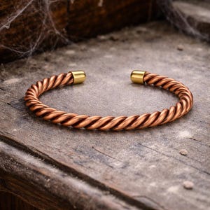 May include: A copper-coloured, twisted metal cuff bracelet with gold-coloured end caps. The bracelet is open and rests on a weathered wooden surface. The twisted design gives the bracelet a rope-like appearance.