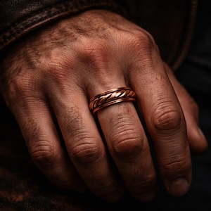 Puede incluir: Un anillo de color cobre con un diseño trenzado. El anillo está en el dedo de una persona. El patrón trenzado del anillo es el punto focal de la imagen. El anillo es una banda simple con una superficie texturizada.