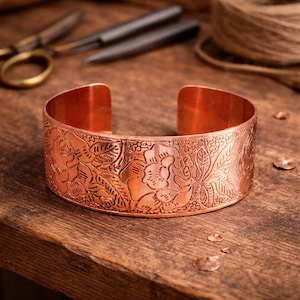 Könnte beinhalten: Ein kupferfarbenes Armband mit geprägtem Blumendesign. Das breite Armband zeigt detaillierte Rosen und Blätter mit einer leicht gebogenen Form. Das Metall hat einen warmen, rötlich-braunen Farbton.