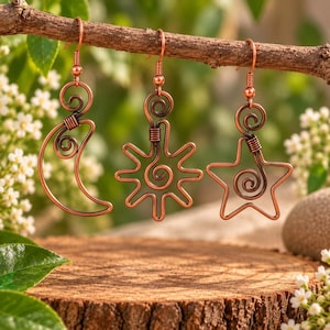 Puede incluir: Tres pendientes de alambre de cobre colgados de una rama, con forma de media luna, un sol con rayos y una estrella de cinco puntas. Cada pendiente está elaborado con alambre de cobre doblado con detalles en espiral y un gancho.