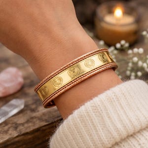 May include: A gold-colored metal bracelet with engraved circular designs and a copper-colored rope-like border. The bracelet is worn on a wrist, and the background includes a lit candle and crystals.