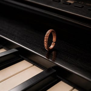 May include: A copper-colored ring featuring a continuous, ribbed, or coil-like texture around the entire band. The ring has a small, visible gap, indicating an adjustable open-ended design, and displays a metallic, reflective finish.