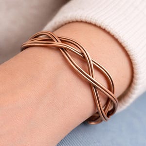 Op de afbeelding: Een koperkleurige armband met een verweven ontwerp. De armband is gemaakt van meerdere strengen metaal die zijn gedraaid en gekruist. De armband wordt om een pols gedragen.