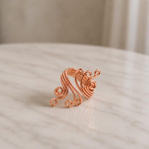 May include: A rose gold-coloured wire ring featuring a series of interconnected, swirling loops that form an open, asymmetrical design. The metal wire is coiled and bent into multiple circular patterns, creating a continuous, flowing structure.