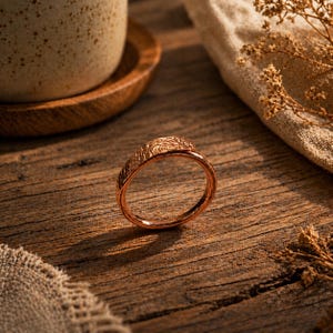 May include: A copper-toned ring with a textured band and a detailed design. The ring sits on a wooden surface, with a cup and saucer in the background. The ring's design has an organic feel. The ring is likely made of copper or a copper alloy.