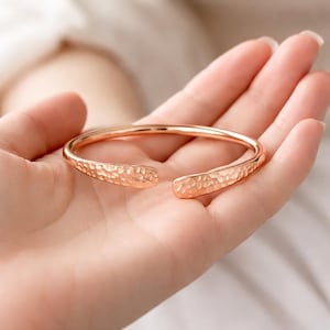 Puede incluir: Un brazalete abierto de oro rosa con una textura martillada. El brazalete está abierto en los extremos y descansa sobre la palma de una persona. La textura martillada le da al brazalete un aspecto único y artesanal.