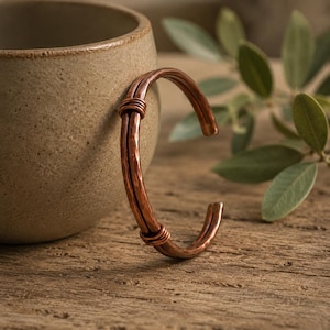 May include: A copper cuff bracelet featuring a hammered texture and two parallel bands held together by two small wire-wrapped sections. The metal has a polished, reddish-brown finish with a curved, open-ended design that maintains a consistent shape.