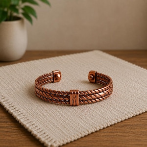 Op de afbeelding: Een koperkleurige manchetarmband bestaande uit drie parallelle gedraaide touwachtige banden die in het midden door een verticale staaf worden verbonden. De armband heeft afgeronde, gladde uiteinden en een metallic afwerking in een roodbruine tint.