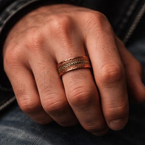 Puede incluir: Un anillo de color cobre con un diseño trenzado y una banda de color dorado. El anillo se lleva en un dedo. El anillo tiene una superficie texturizada y una banda de metal dorado.