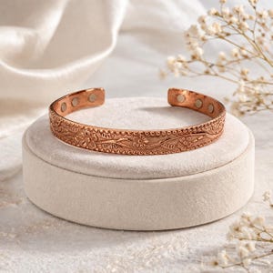 May include: A copper-coloured cuff bracelet with a floral pattern and magnetic closures. The open-ended bracelet is displayed on a white, round pedestal. The bracelet's design features detailed floral and leaf motifs.