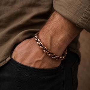Op de afbeelding: Een koperkleurige kettingarmband met een gevlochten ontwerp. De armband wordt om een pols gedragen, met een bruin overhemd en een zwarte jeans op de achtergrond. De armband is een opvallend accessoire.