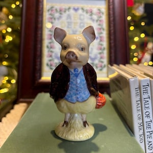 May include: A ceramic pig figurine, dressed in a blue waistcoat and brown jacket, carrying a red bag. The pig stands on a grassy base. It's positioned on a green book, with a framed embroidery visible in the background.