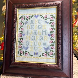May include: A framed cross-stitch sampler featuring the alphabet, flowers, and birds. The embroidery uses pastel colours, framed in a dark wood frame with ornate detailing. The background is a light cream colour.