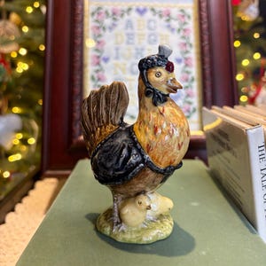 May include: A ceramic hen figurine with two chicks. The hen is wearing a black bonnet and a black shawl. The hen has brown and orange feathers. The chicks are yellow. The figurine is on a green surface.