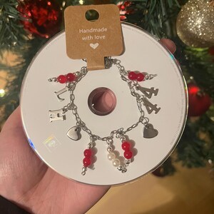 Peut inclure: Un bracelet fait à la main avec des maillons de chaîne argentés, des perles rouges et des breloques en forme de cœur. Le bracelet est présenté sur un CD et porte les lettres "L" et "H" en breloques. Une étiquette indique "Handmade with love".