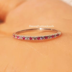 May include: A thin silver-toned band ring featuring a row of alternating small round red and clear stones set along the top half. The text HansrajFashionJewels is printed in small white letters above the center of the ring band.