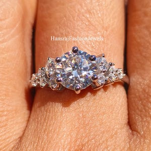 Könnte beinhalten: Ein silberner Verlobungsring mit einem großen, runden, klaren Mittelstein und kleineren Akzentsteinen. Der Ring ist mit mehreren Krappen gefasst und hat ein zartes Band. Der Text "HansrajFashionJewels" ist sichtbar.