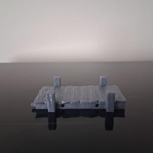 May include: A small, gray, 3D-printed miniature dock platform featuring a textured wooden plank surface and four vertical corner posts. The rectangular structure has a weathered appearance with visible grain details on the planks and a solid base.