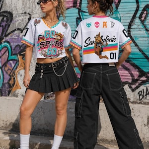 May include: Two people wearing white mesh crop tops with colorful graphics and the text "GO SPURS GO" and "San Antonio". The tops have black numbers and are paired with black bottoms and sneakers. The background features graffiti art.