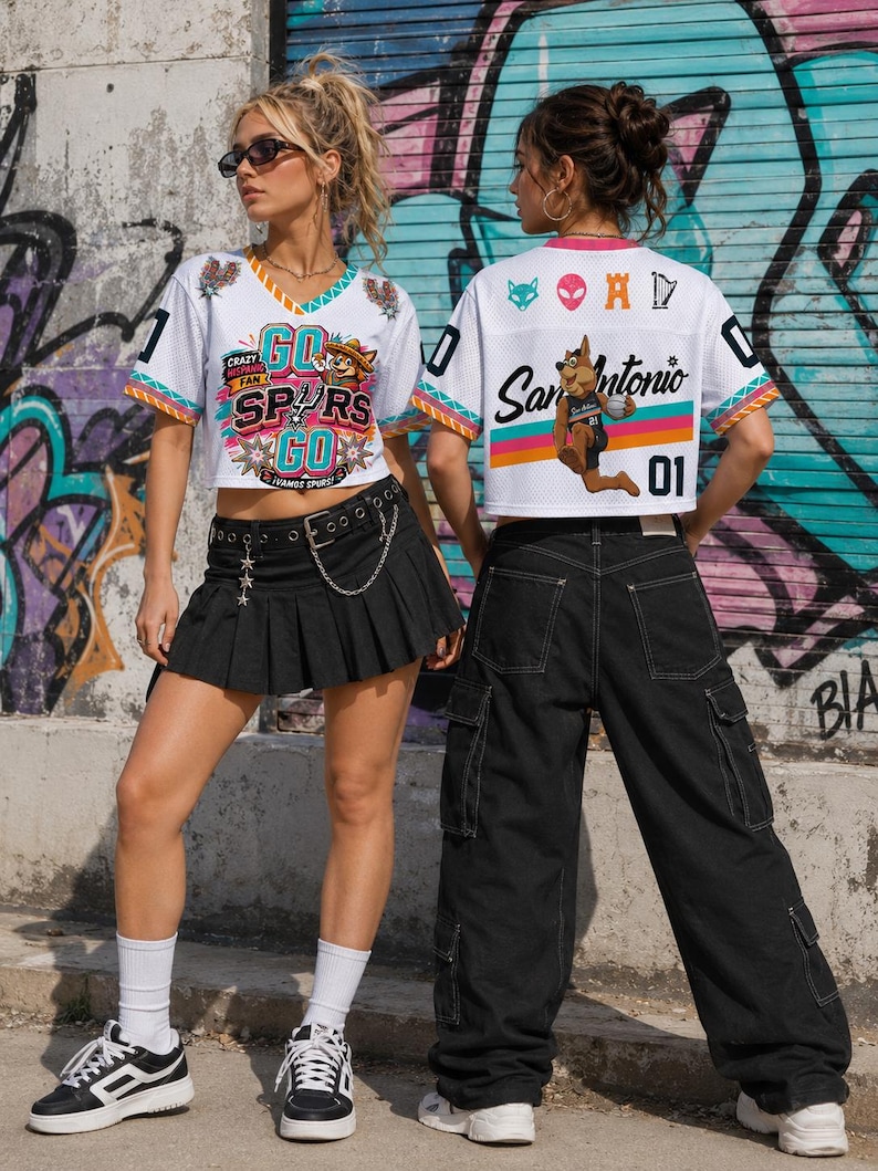 May include: Two people wearing white mesh crop tops with colorful graphics and the text "GO SPURS GO" and "San Antonio". The tops have black numbers and are paired with black bottoms and sneakers. The background features graffiti art.