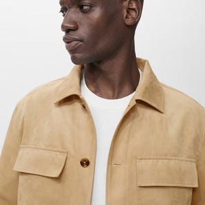 May include: A tan suede jacket with two front pockets and button closures. The jacket is worn over a white t-shirt. The model has short, dark hair and is looking to the side. The background is plain white.