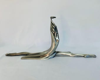May include: A silver-toned sculpture depicting a stylized human figure in a graceful pose. The sculpture features elongated limbs and a flowing, abstract design, suggesting movement and fluidity. The artwork is set against a plain white background.